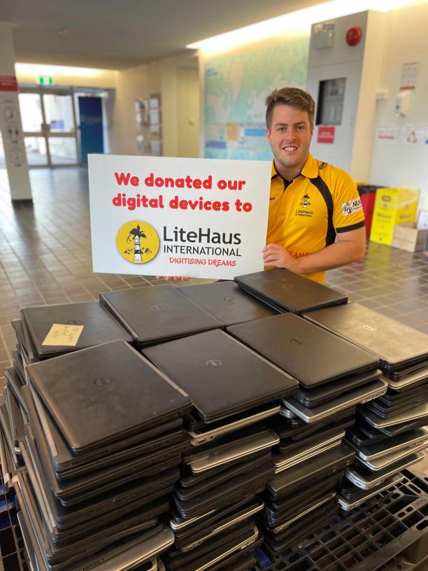  Alumni Jack Growden holds up a sign saying 'we donated our digital devices to LiteHaus International' while he smiles standing behind a large pile of laptops on a desk. 