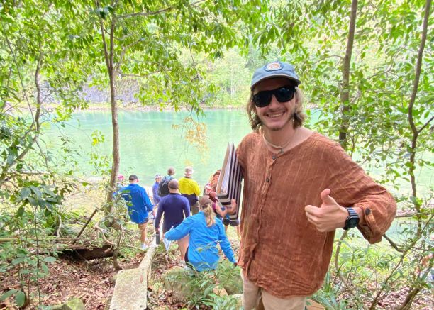  student Indus Fisher smiling with thumbs up in a rainforest with students in the background at a creek. 